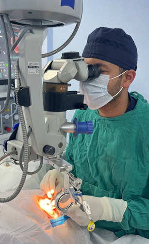 Surgeon in green scrubs and a blue cap operating with a surgical microscope on a patient’s eye in a sterile operating room.