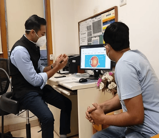 Doctor wearing a mask and patient discuss eye health at a clinic; monitor shows an eye diagram on screen